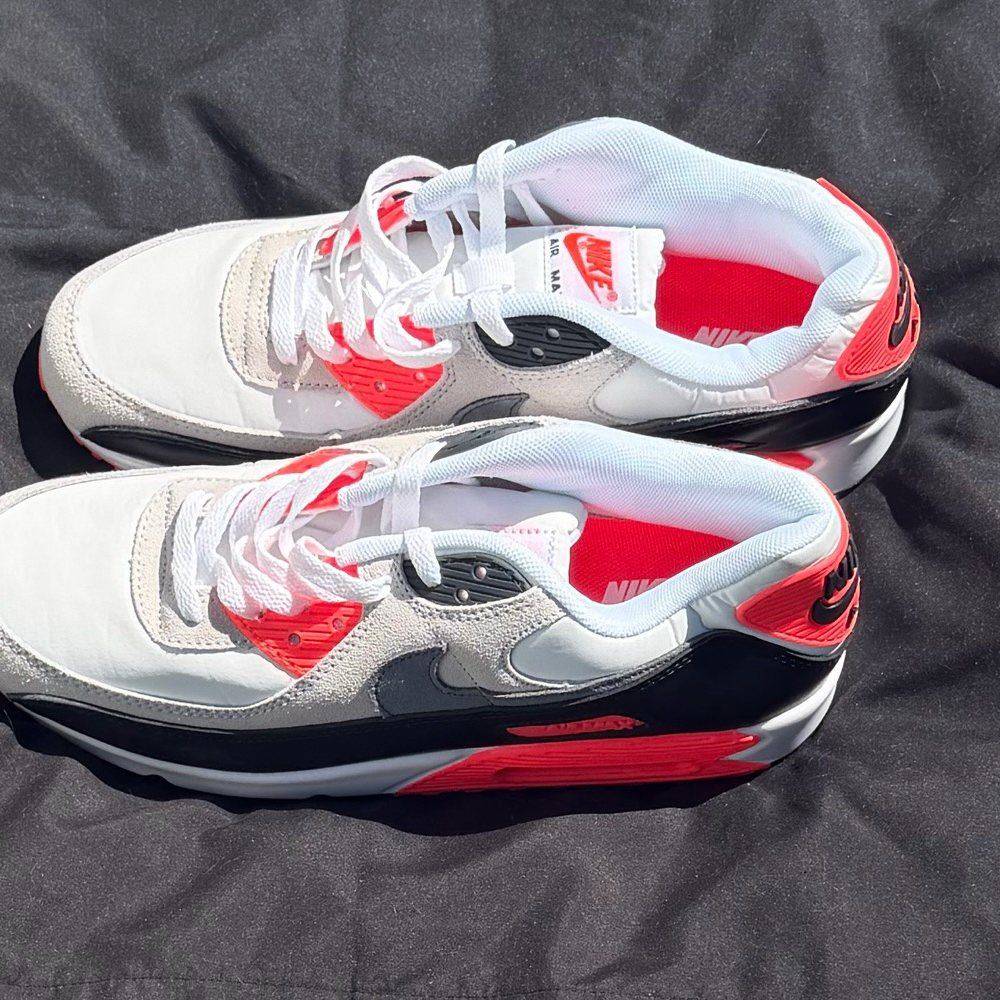 A pair of white, gray and black Nike Air Max 90 sneakers with bright red trim, laid out on a dark cloth